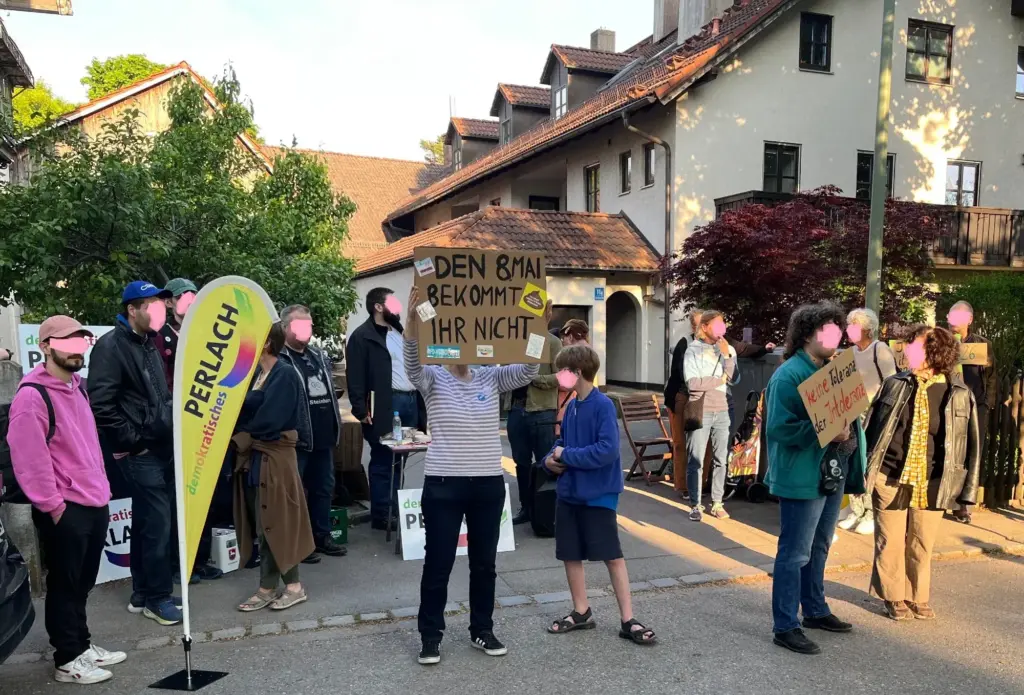 Menschen stehen auf der Straße zusammen. Einige halten selbstgemalte Schilder hoch. Dort steht: "Keine Toleranz der Intoleranz" und "Den 08. Mai bekommt ihr nicht". Zu sehen ist auch noch ein Beachflag mit dem Logo von demokratisches Perlach.