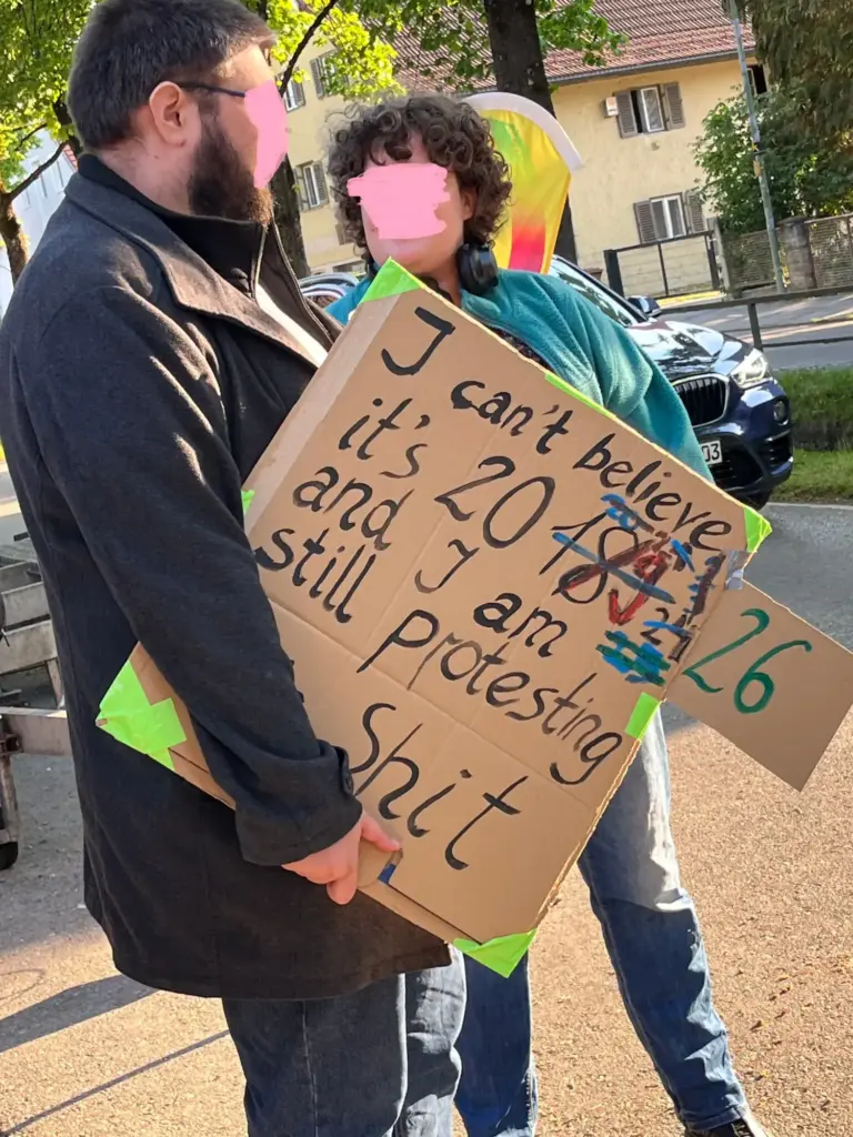 Zwei Personen - Ein Mann und eine Frau- in Nahaufnahme. Der Mann hat ein selbstgemaltes Schild under dem rechten Arm. Dort steht: I can´t believe, it´s 20.... (und hier sind viele durchgestrichene Zahlen zu sehen: 18, 19, 23, 24...) und auf einem zusätzlich angeklebten, herausragendem kleinen Stück Pappe steht die Zahl 26. Dann geht der Text auf dem großen Schild weiter: ...and I am still protesting this shit.