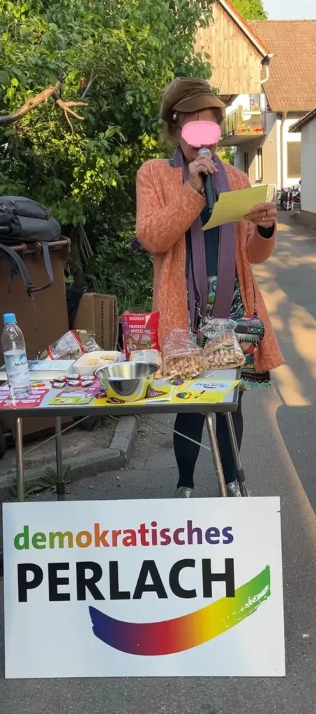 Eine Rednerin mit Mikrofon steht hinter einem kleinen Tisch (mit Essen und Flyern). Sie hält ihr Manuskript hoch und spricht in das Mikro. Vor dem Tisch steht ein Schild mit: "demokratisches Perlach"-Logo in bunter Farbe auf der Erde. Im Hintergrund ist ein Baum, eine Einfahrt und ein Haus zu sehen.
