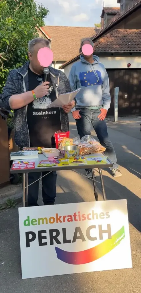 Ein Redner steht hinter einem kleinen Tisch (mit Essen und Flyern darauf). Er hält ein Mikro in der Hand, spricht rein und in der anderen Hand hat er sein Mauskript. Im Hintergrund steht ein weiterer Mann mit einer EU-Flagge auf seinem Sweater abgebildet. Vor dem Tisch auf der Erde steht das Schild mit "demokratisches Perlach"-Logo in bunten Farben. Im Hintergrund ist ein Baum, eine Einfahrt und ein Haus zu sehen.